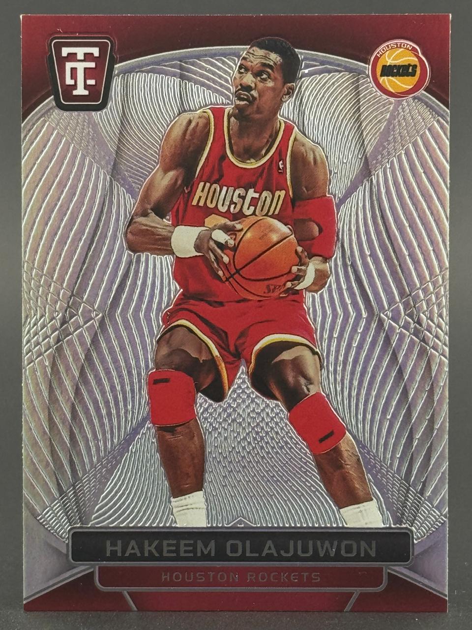 Hakeem Olajuwon trading card with silver textured background holding a basketball in a Houston Rockets uniform.